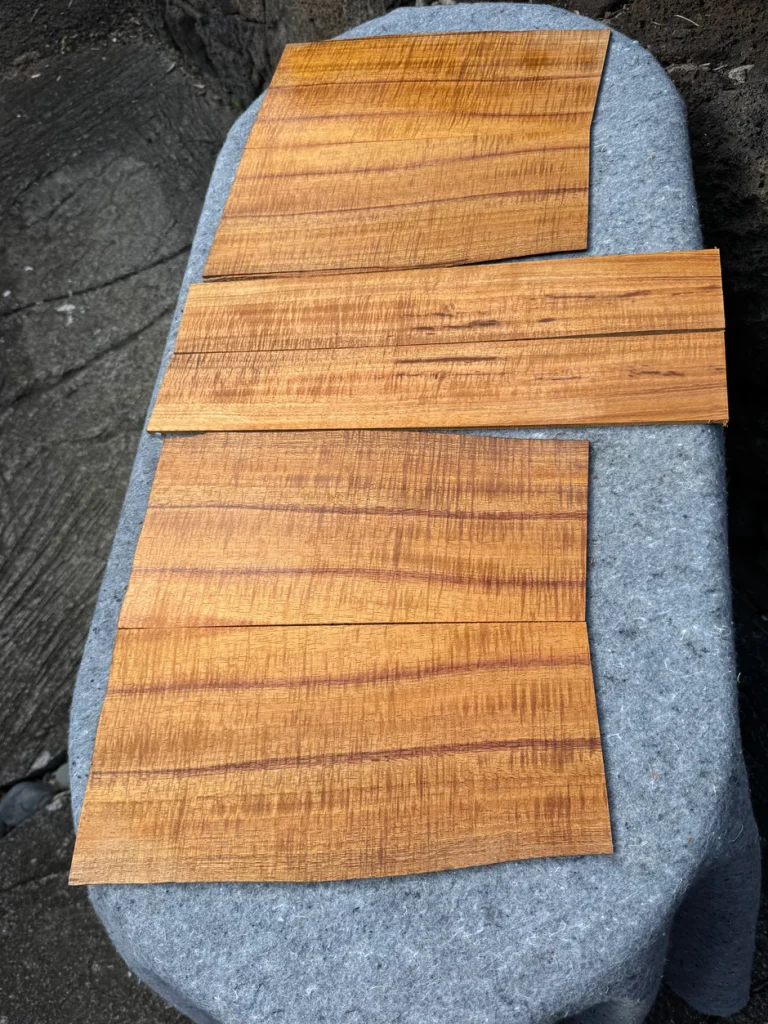 Three thin rectangular wood veneer panels with warm golden-brown tones and visible wavy grain patterns laid flat on a gray fabric surface outdoors.